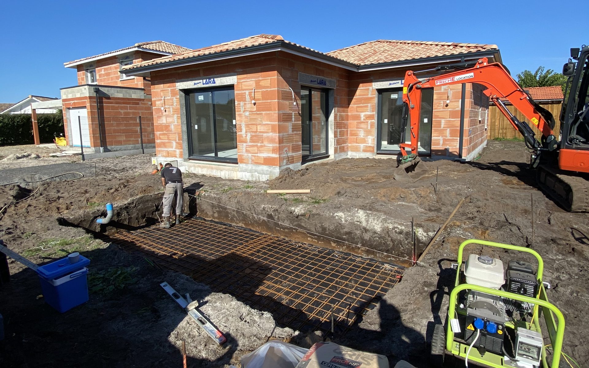 Chantier de terrassement et ferraillage de fondations avec mini-pelle devant une villa en briques